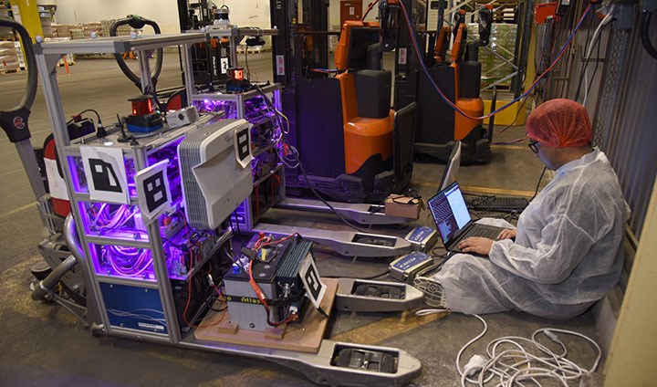 En forskare sitter på golvet bakom två truckar. Han har skyddskläder och tittar intensivt på en dator.