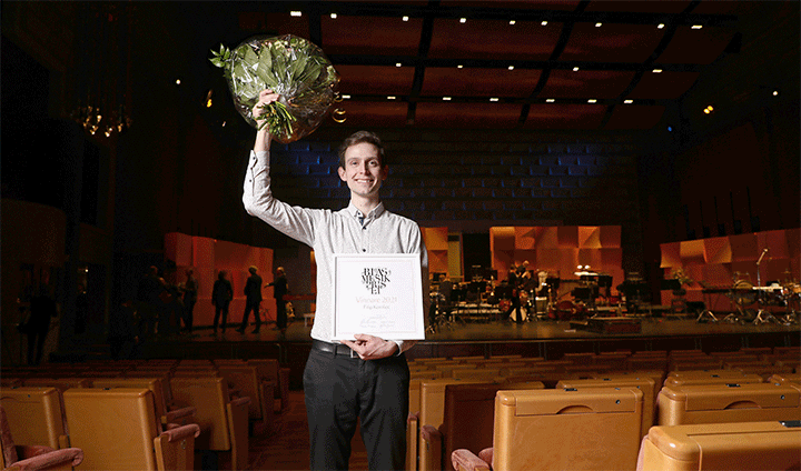 Man standing in a dark music hall with an orchestra on stage behind him and empty chairs around the hall. With his left hand, he is holding a diploma. In his right hand, he is holding a flower bouquet above his head.