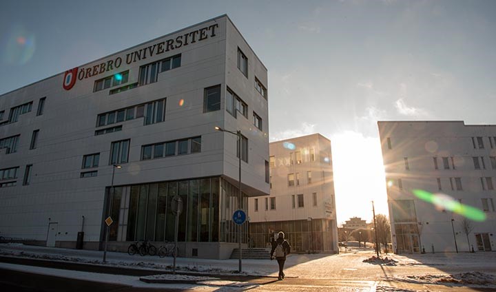 Solen tittar fram bakom Örebro universitets vita byggnader. I förgrunden syns Novahuset, med en stor skylt på fasaden där det står Örebro universitet.