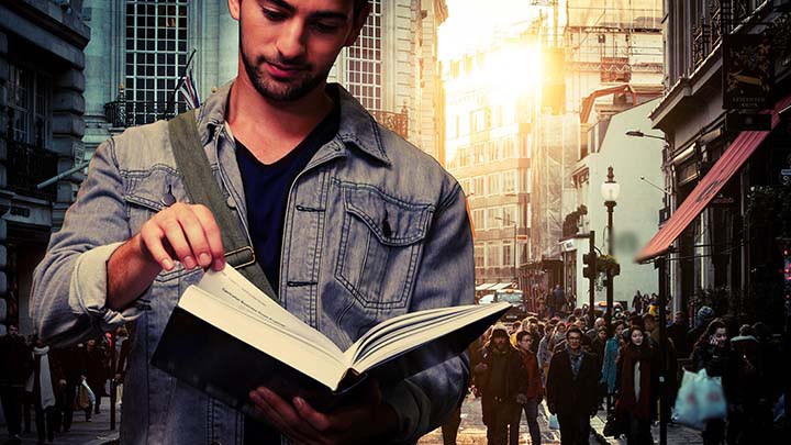Foto på en person som står och läser i en bok med en folkrik stadsgata i bakgrunden.