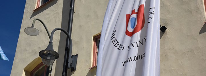 Örebro universitets logga på en vit beachflagga. Beachflaggan står framför en husvägg av sten. 