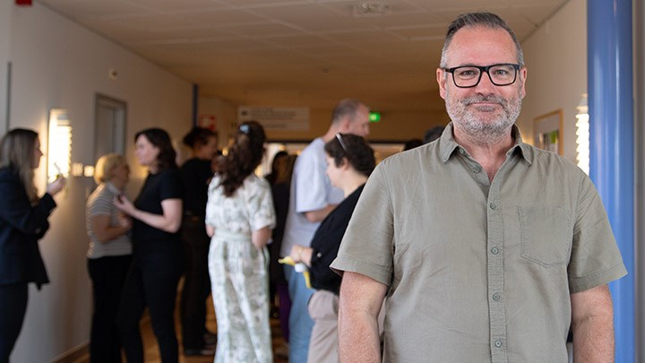 A man with a gray-green shirt and glasses. In the background, there is a group of people.