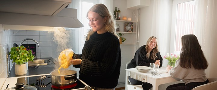 Tre studenter i ett kök där en student kokar pasta och de andra sitter vid ett bord.