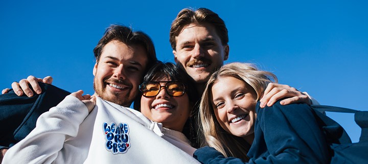 Fyra studenter håller om varandra under klarblå himmel. En av studenterna visar upp ett orubros-märke som sitter fast på tröjan. 