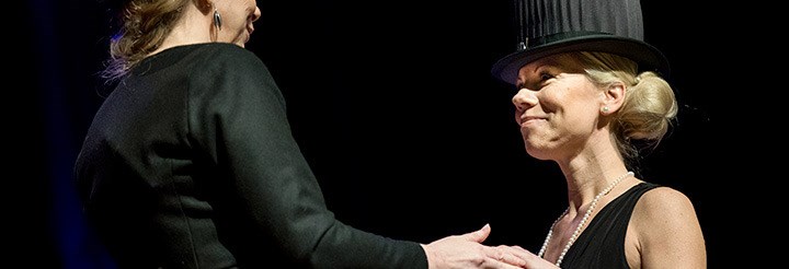 Two women in black clothes and black doctor's hats are holding hands. They look at each other.