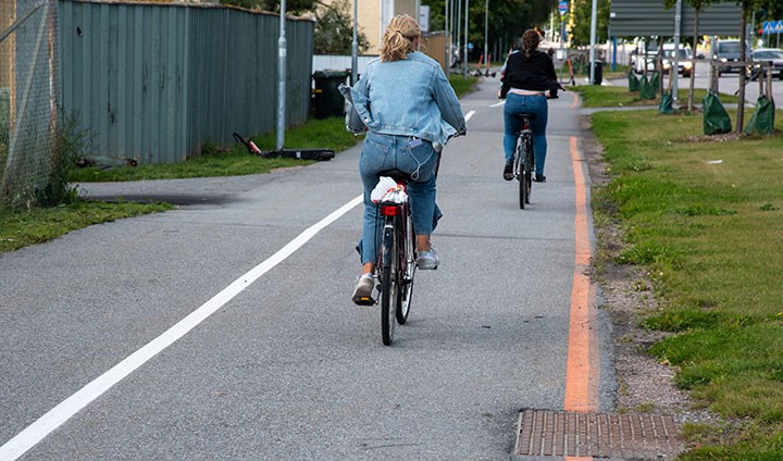 Två cyklister cyklar på en asfalterad cykelbana, ett huvudcykelstråk i Örebro. I kanten av cykelbanan är ett orange streck målat.