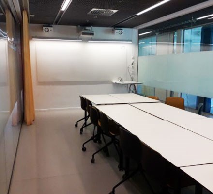 Vy 2 - Rektangulära bord med stolar runt framför en whiteboardtavla. Glasvägg vid sidan.