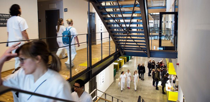 Female student wearing white lab clothes walking upstairs, with a group of students wearing white lab clothes in the lobby below.