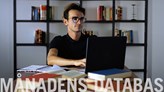 Person som sitter vid ett skrivbord med en dator och flera böcker. I förgrunden står det "Månadens databas". 
