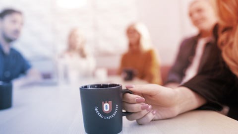 Fem personer sitter runt ett bord. I förgrunden håller en person i en mugg marinblå mugg med Örebro universitets logga.