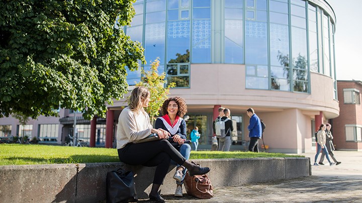 Två studenter sitter och pratar utomhus framför Universitetsbiblioteket.