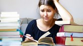 Foto på en person som stressad tittar i en bok, omgiven av travar med böcker.