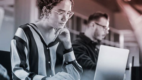 A woman is viewing a computer with a man in the background.