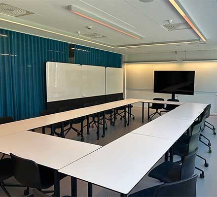 Vy 2 - Rektangulära bord och stolar ihopställda i en rektangel. Rullbara whiteboardtavlor. Gardiner på en vägg.