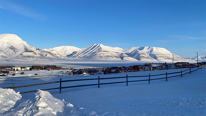 A winter landscape showing a fence and snow-covered mountains with a blue sky above.