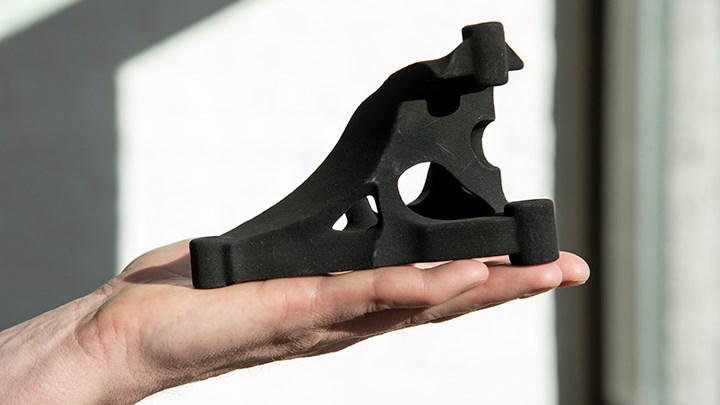 En hand håller upp en 3D-printad svart fordonsdel. Den fyller hela handflatan, men är lätt att lyfta.