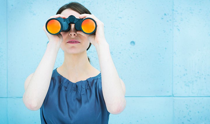 A woman is standing looking in a binoculars. She has a blue top.