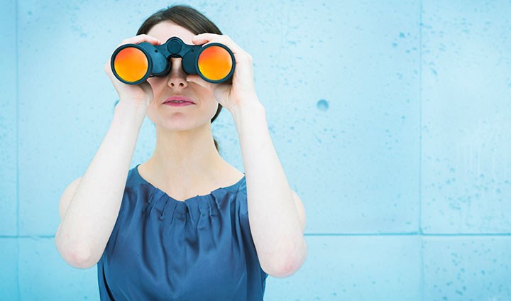 A woman is standing looking in a binoculars. She has a blue top.