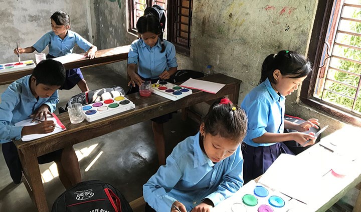 Fyra tjejer och en kille sitter i ett klassrum och målar med vattenfärger. Alla har blå skoluniformer på sig.