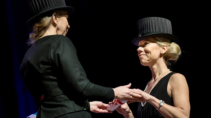 Två kvinnor i svarta kläder och svarta doktorshattar håller varandra i händerna. De tittar på varandra.