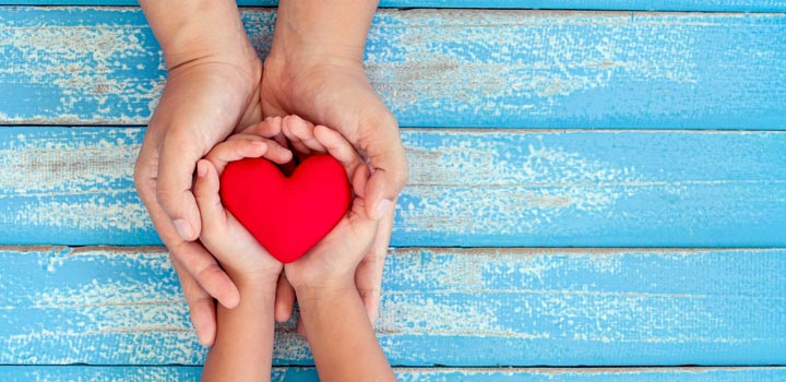 A red heart lying in cupped hands of a child, hands which in turn rests in hands of an adult
