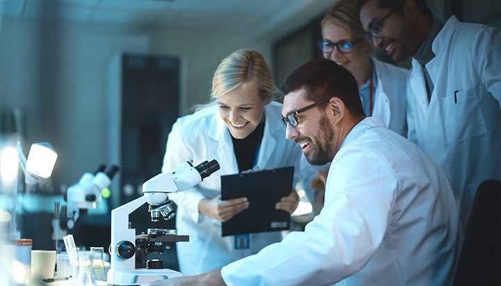 A microscope in the foreground, in the background four scientists in discussion.