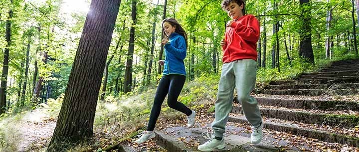 Två barn springer i ett löpspår i skogen med sol i bakgrunden mellan träden