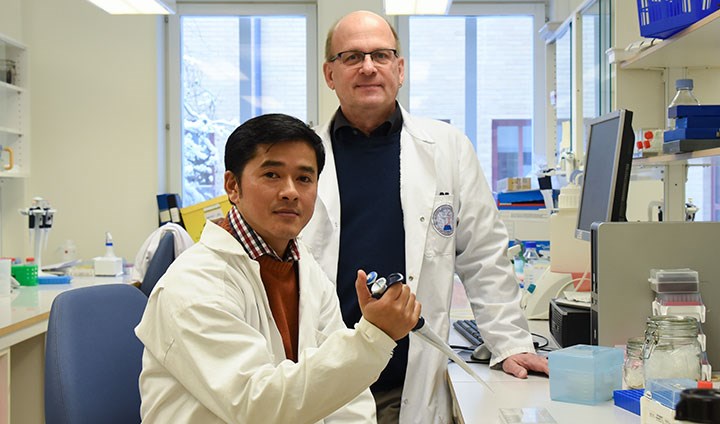 Two men in white lab coats, one sitting and holding a test instrument, the other standing behind