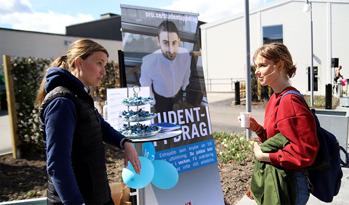 Två personer pratar. En skylt där det står Studentuppdrag syns i bakgrunden.