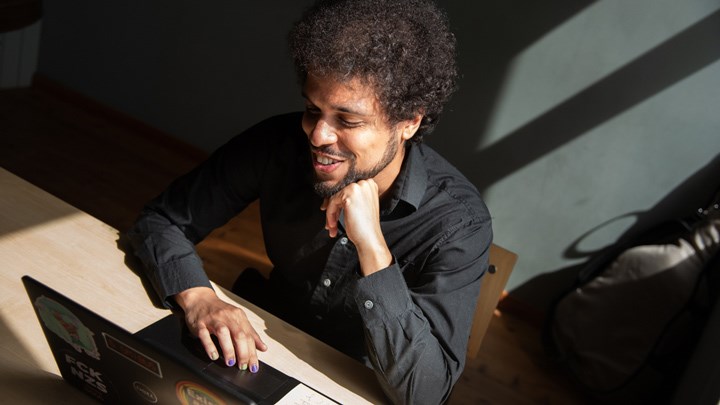 A person working on a laptop while seated at a table in a sunlit room.