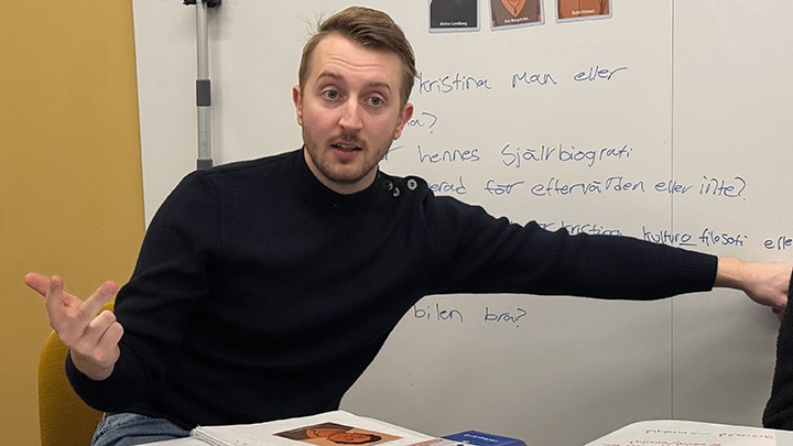 A student talks and counts on his fingers in front of a whiteboard.