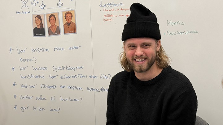A student wearing a cap smiles in front of a whiteboard.