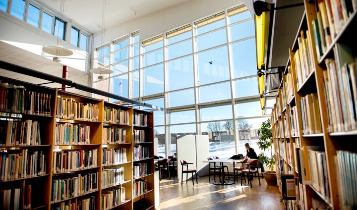 Invändig bild från biblioteket med bokhyllor i förgrunden och stora glasfönster i bakgrunden.