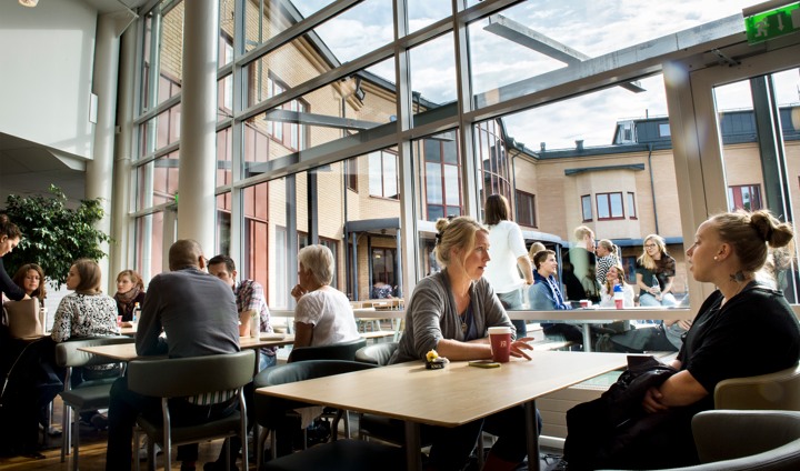 Studenter vid bord och stora glasfönster i bakgrunden.