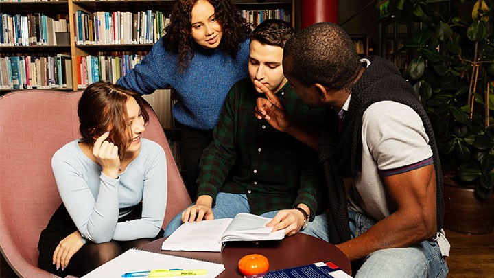Fyra studenter diskuterar runt ett bord framför en bokhylla
