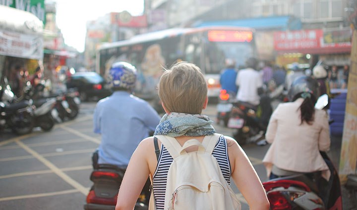 En person med ryggsäck syns bakifrån tittandet ut över en gatumiljö som inte ser svensk ut