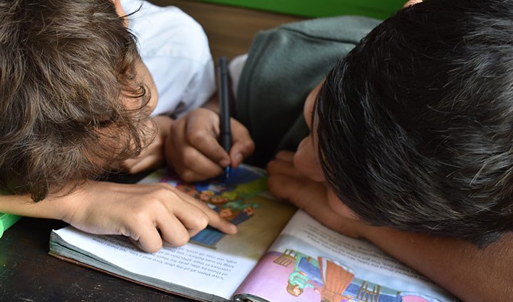 Två barn siter med huvudena ihop och skriver och läser i en bok.