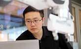 Researcher Da Sun, sitting in the robotics lab beside three robot arms.