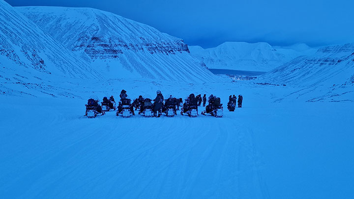Students riding snowmobiles towards snow-covered mountains.