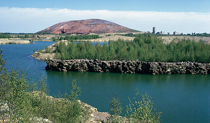 Photo of water-filled open pits with plant life on land. Kvarntorp, with its hill and industrial area, is in the background.